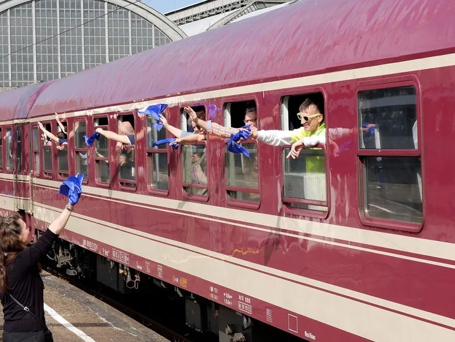 Abschied: Aus einem Zugwagon winken mehrere Männer mit blauen Tüchern aus offenen Fenstern. Eine Frau (ebenfalls mit blauem Tuch) winkt zurück