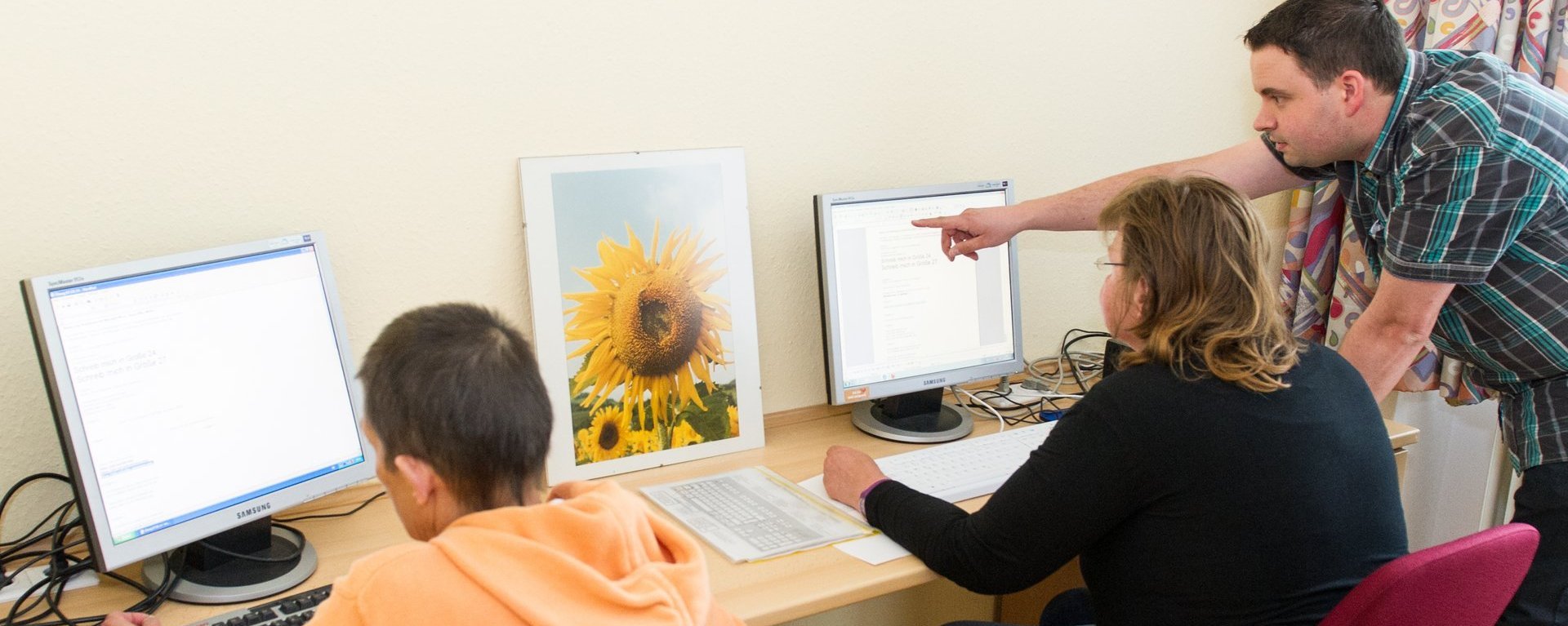 Computer-Unterricht: zwei Männer sitzen an Computern und üben Textverarbeitung. Ein Lehrer gibt Anweisungen und zeigt dabei auf den Monitor