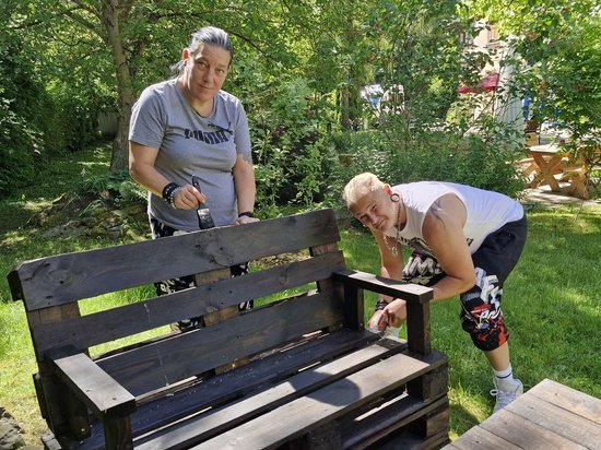 Zwei Personen streichen eine aus Holzpaletten gebaute Gartenbank im Freien dunkelbraun. Eine Frau und ein Mann arbeiten gemeinsam an dem selbstgebastelten Möbelstück.