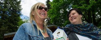 In der Natur: Zwei Frauen lachen. Eine hält ein kleines Vogelhäuschen in der Hand