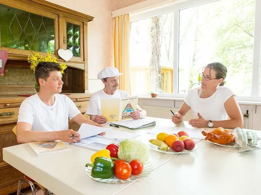 Die Küchen-Crew bei der Menüplanung: eine Frau und zwei Männer (einer mit Kochmütze und Ernährungsbroschüre) sitzen am Tisch mit diversen Zutaten.