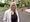 Eine Frau mit langen blonden Haaren steht im Freien. Sie trägt eine helle Jacke über einem schwarzen Oberteil und blauen Jeans. Im Hintergrund sind Bäume und ein gepflasterter Bereich sichtbar.