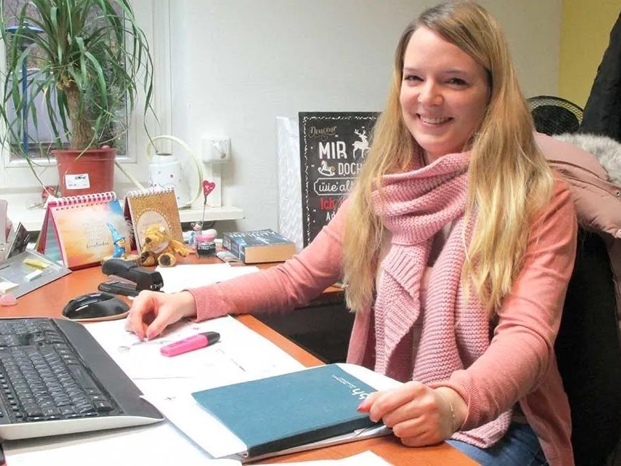 im Büro: eine junge Frau mit langem blonden Haar sitzt an ihrem Arbeitsplatz am Computer und lächelt freundlich in die Kamera