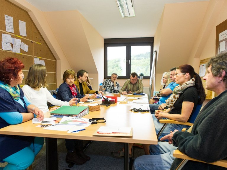 Eine große Gruppe Frauen und Männer sitzen an einem großen Besprechungstisch auf dem sich viele Unterlagen und Büroartikel befinden. Die Mimik zeigen: es wird ernsthaft gearbeitet, gesprochen und nachgedacht.