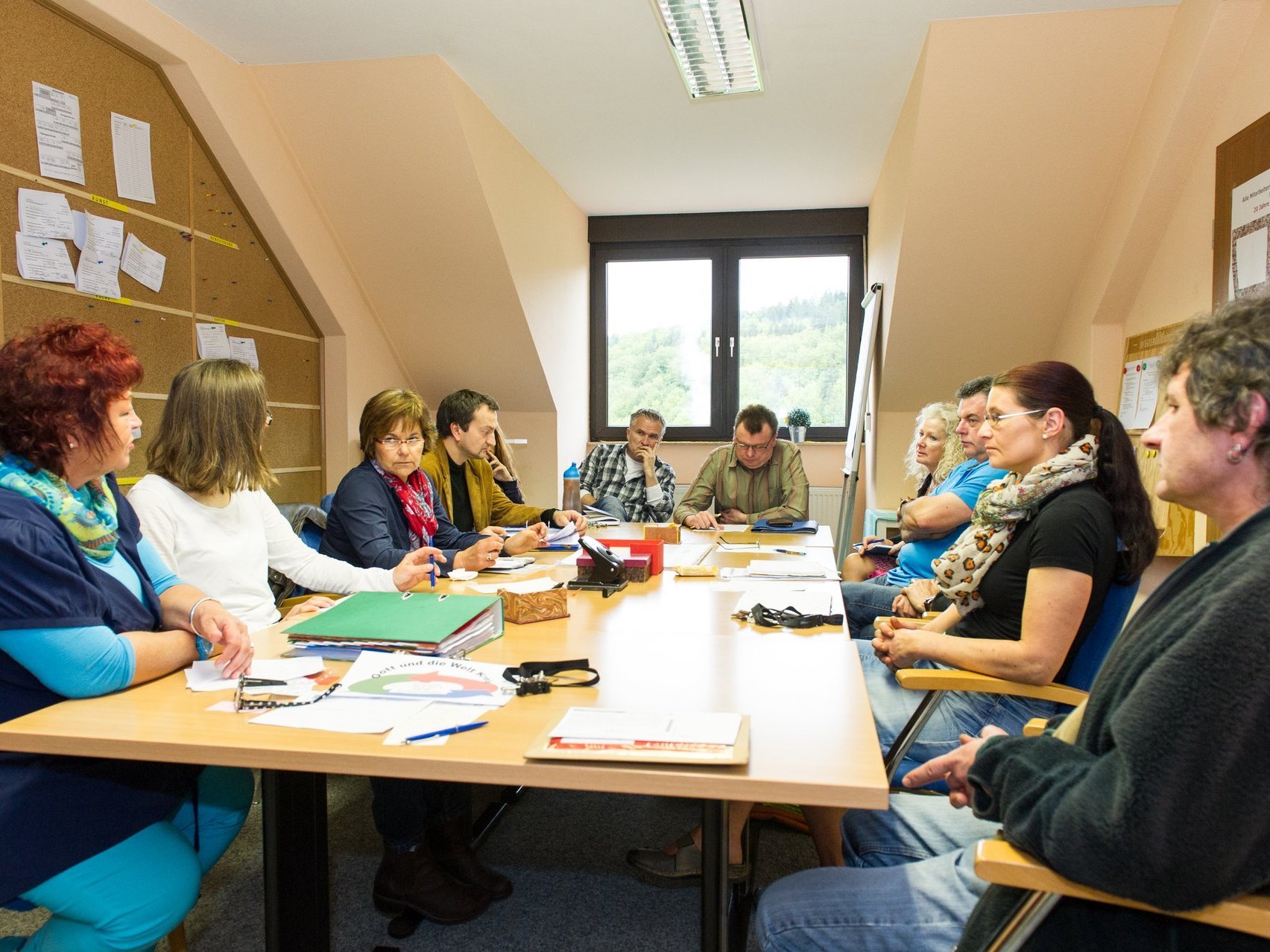 Eine große Gruppe Frauen und Männer sitzen an einem großen Besprechungstisch auf dem sich viele Unterlagen und Büroartikel befinden. Die Mimik zeigen: es wird ernsthaft gearbeitet, gesprochen und nachgedacht.