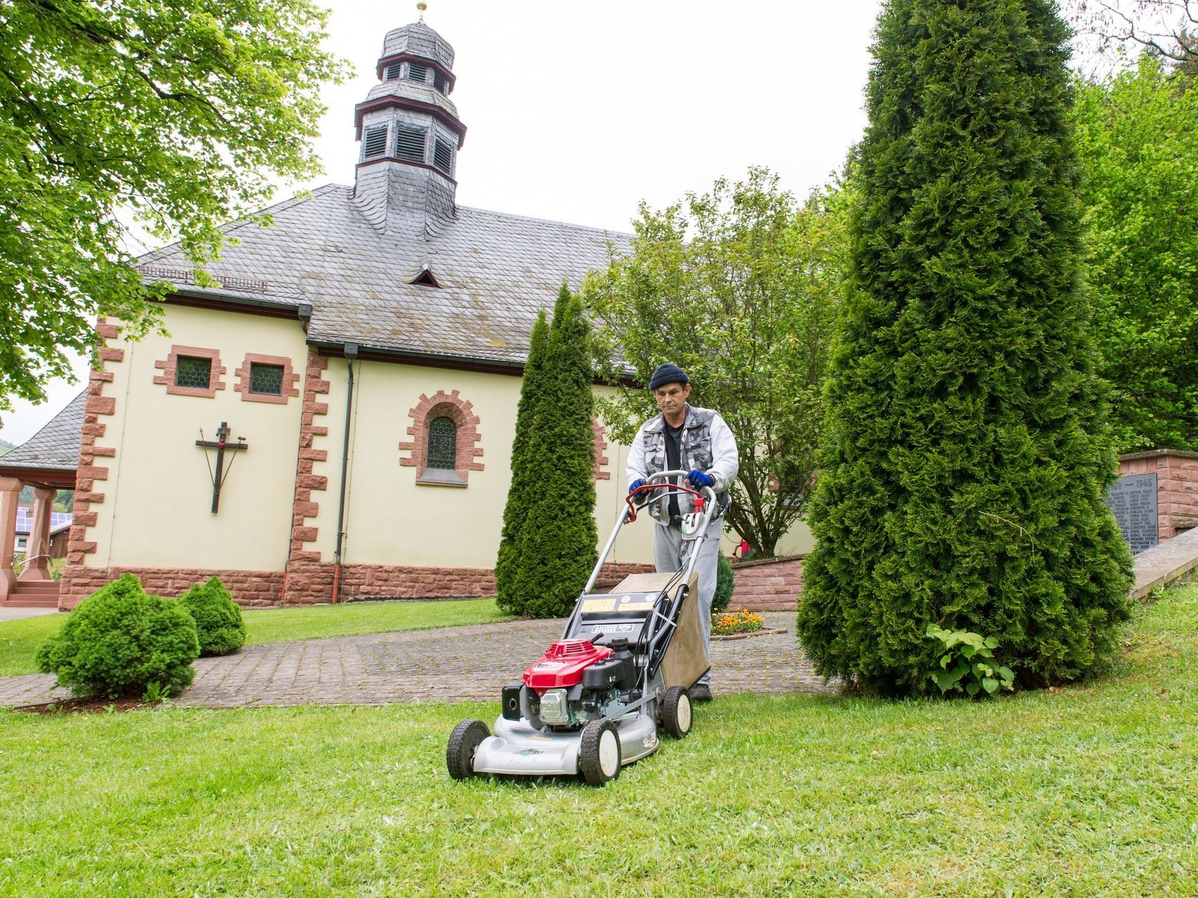 Gartenarbeiten: ein Mann mäht den Rasen vor einer Kapelle