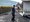 Ein lächelnder Arbeiter in Arbeitskleidung steht auf frisch verlegtem Asphalt und hält ein Werkzeug. Im Hintergrund ist ein weiterer Kollege bei der Straßenbauarbeit zu sehen.