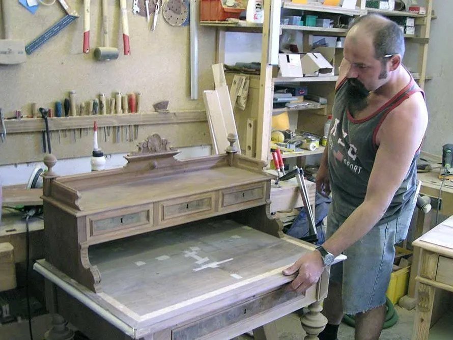 Werkstatt: ein Mann mit langen Kinnbart und Bleistift hinter das Ohr geklemmt restauriert eine alte Kommode