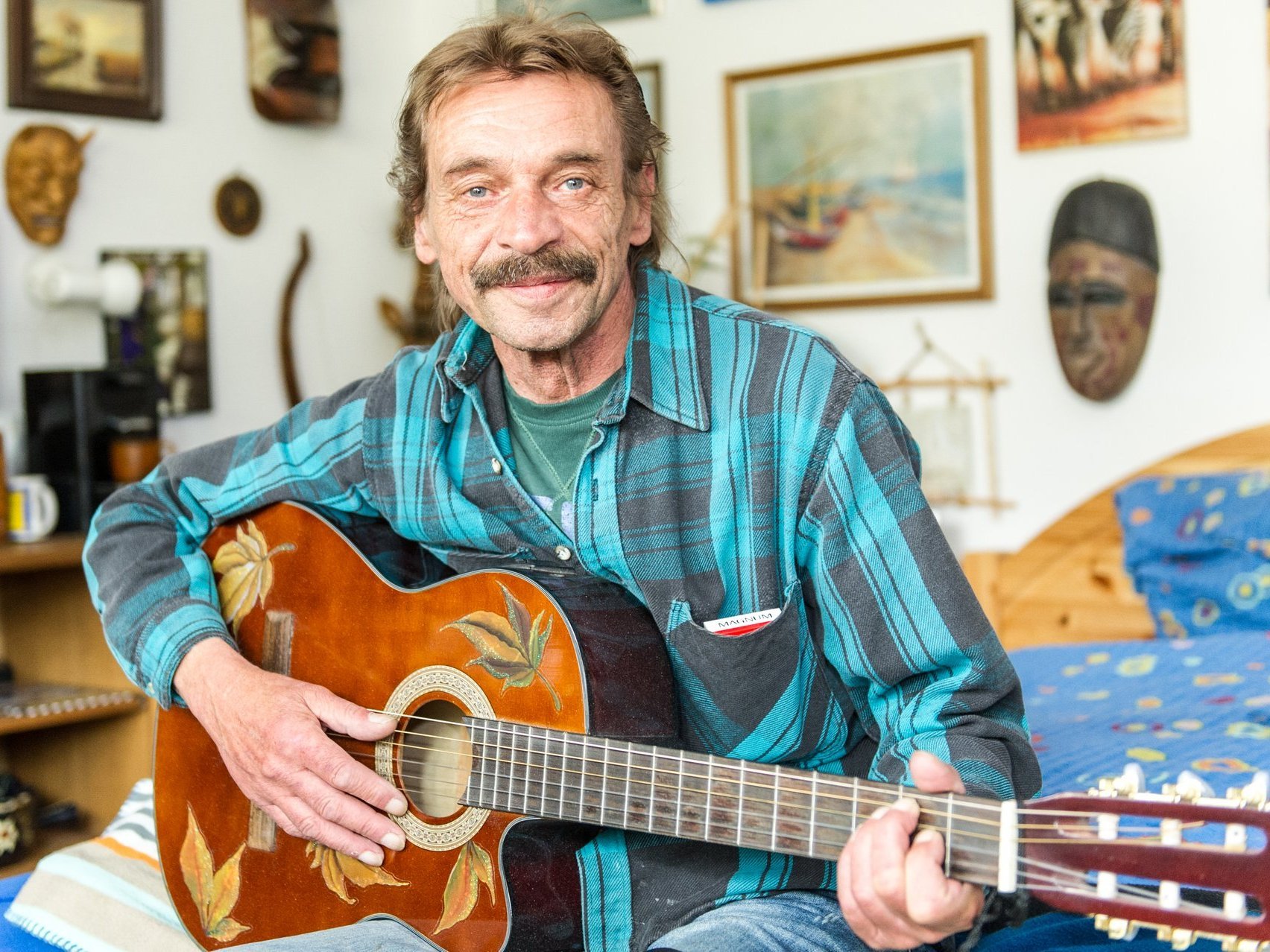 Ein freundlich blickender älterer Mann mit kariertem Hemd spielt auf dem Bett sitzend Gitarre