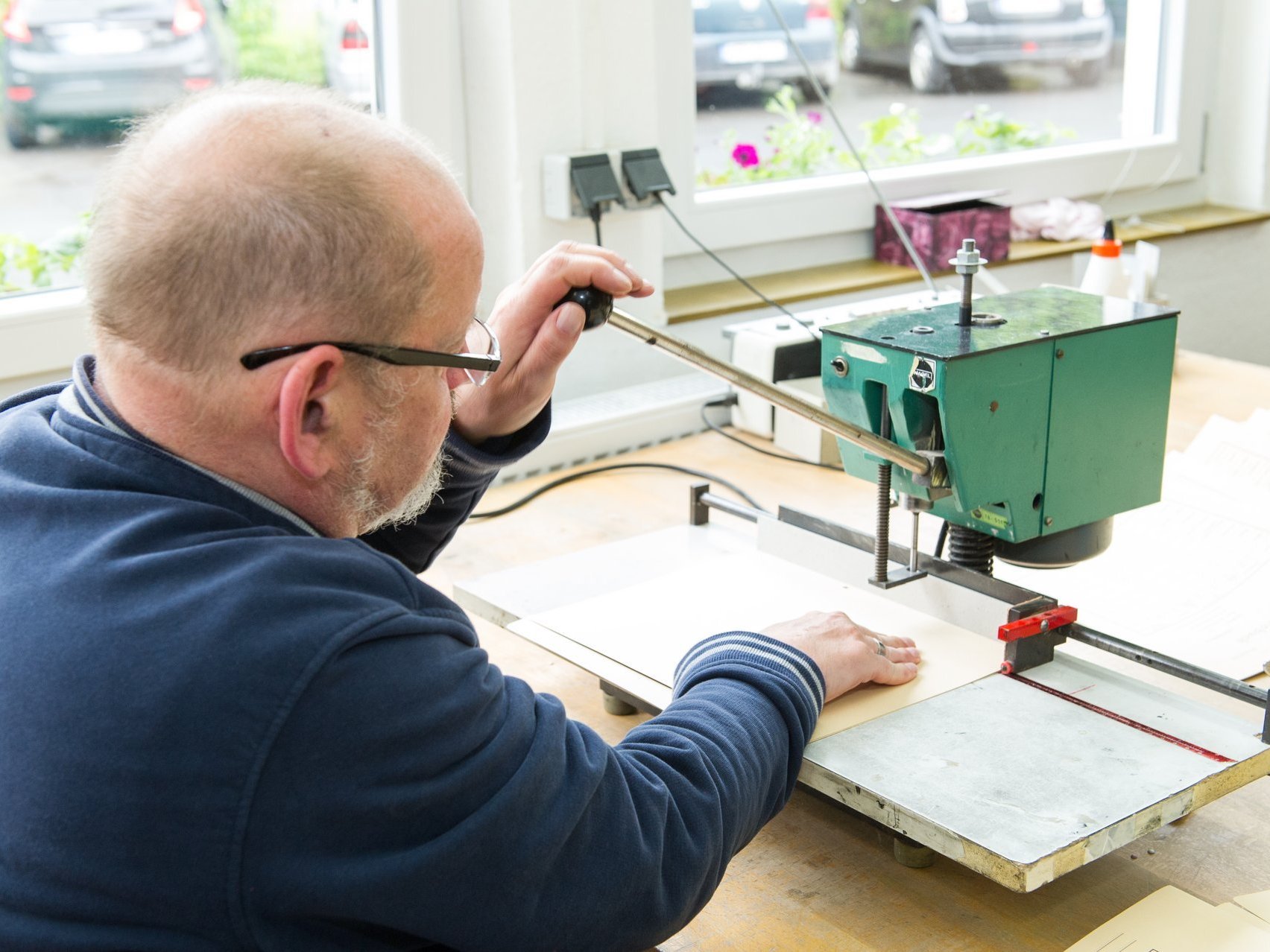 Ein Mann mit grauem Haar und Brille schneidet Papierbögen mit einer Schneidemaschine