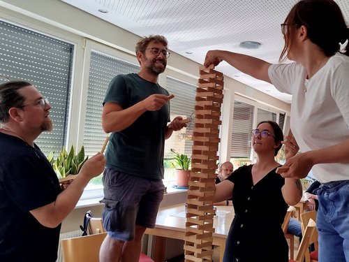 Vier Personen spielen ein Spiel mit Holzklötzen, bei dem ein hoher Turm gebaut wird. Eine Frau greift nach einem Klotz, während ein Mann darauf zeigt. Die Gruppe wirkt konzentriert und amüsiert.
