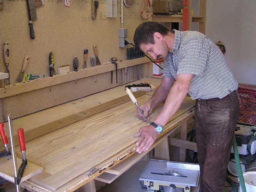 Werkstatt: Ein Mann mit schnurrbart und kurzem Haar markiert mit einem Stemmeisen eine Stelle auf einem dicken Holzbrett