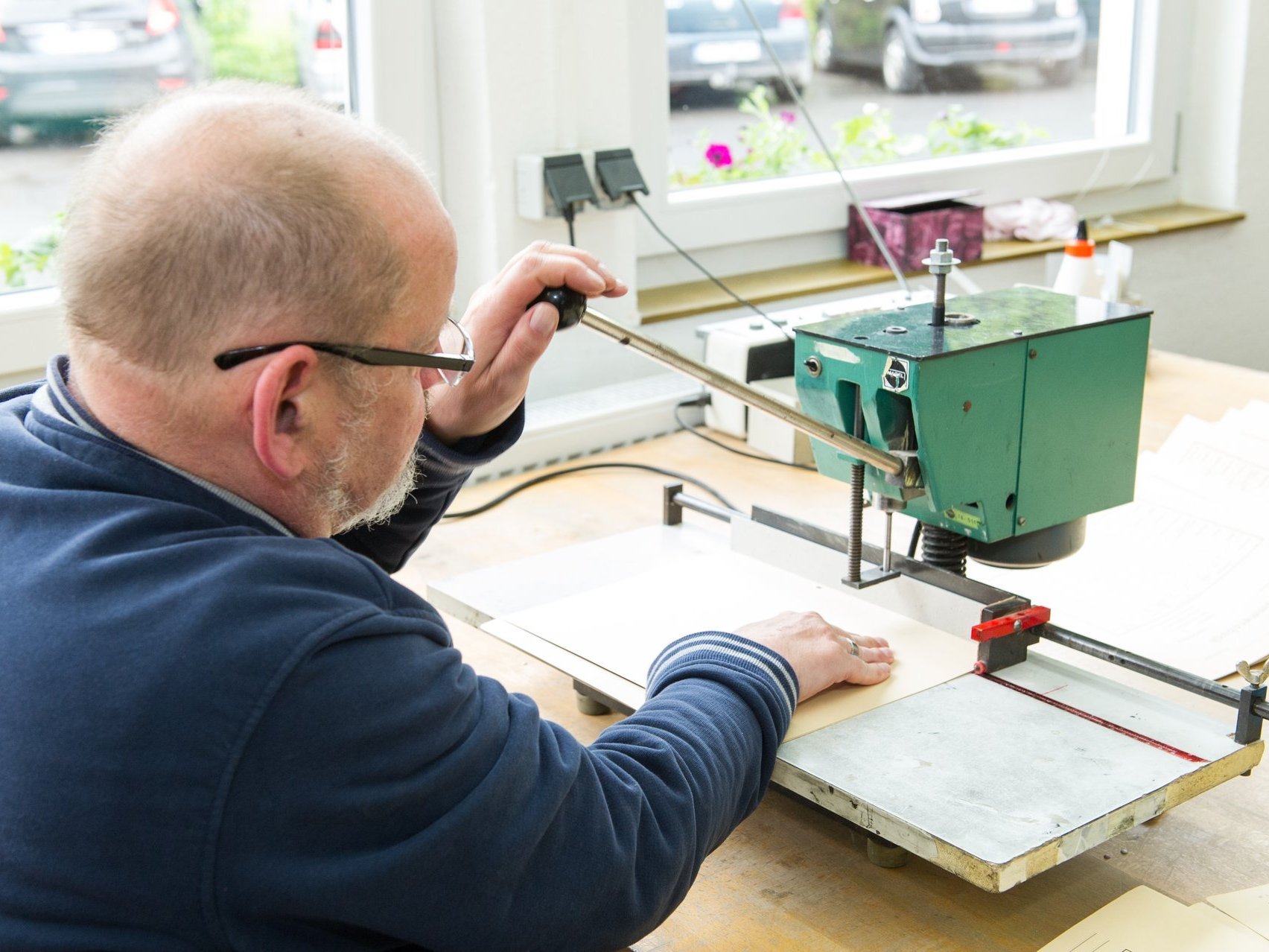 Ein Mann mit grauem Haar und Brille schneidet Papierbögen mit einer Schneidemaschine