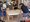 Eine Gruppe spielt ein Teambuilding-Spiel. Vier Personen halten an Seilen, um farbige Becher auf einem Tisch zu bewegen. Zwei weitere Personen beobachten das Geschehen in einem hellen Raum, der wie ein Klassenzimmer oder eine Werkstatt wirkt.