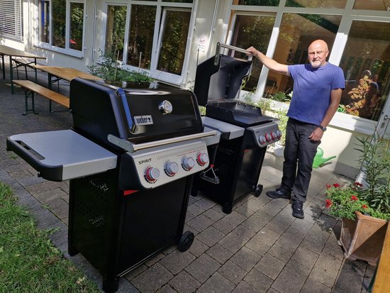 Ein Mann in blauem T-Shirt steht auf einer Terrasse neben zwei Gasgrills und hebt den Deckel des hinteren Grills an, während der vordere Grill geschlossen ist.