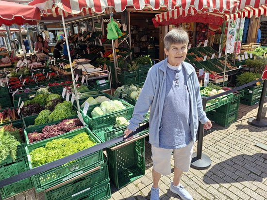 Eine Frau mit kurzen grauen Haaren steht lächelnd vor einem Gemüsestand. Sie trägt eine hellblaue Jacke und Shorts. Im Hintergrund sind grüne Kisten mit frischem Gemüse unter einem rot-weiß gestreiften Vordach zu sehen.