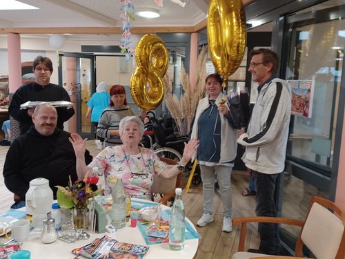 An einem Tisch feiert eine ältere Dame ihren 80. Geburtstag und winkt fröhlich. Ein Mann hält goldene „80"-Ballons hoch, während eine Frau mit einem Mikrofon steht. Ein älterer Herr sitzt neben ihr. Im Hintergrund ist ein Gemeinschaftsraum mit weiteren Personen zu sehen.