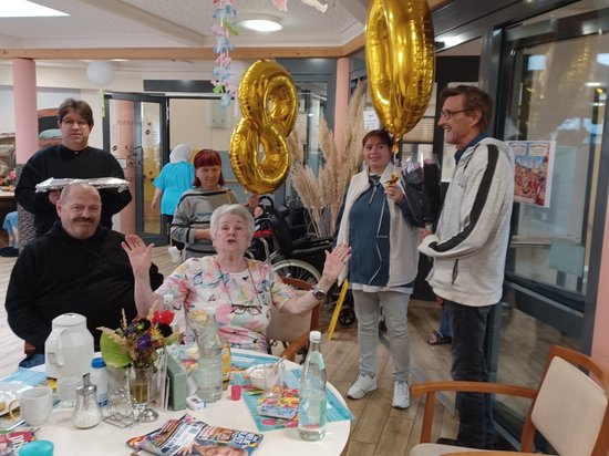 An einem Tisch feiert eine ältere Dame ihren 80. Geburtstag und winkt fröhlich. Ein Mann hält goldene „80"-Ballons hoch, während eine Frau mit einem Mikrofon steht. Ein älterer Herr sitzt neben ihr. Im Hintergrund ist ein Gemeinschaftsraum mit weiteren Personen zu sehen.