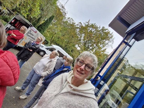 Eine lächelnde Frau mit kurzen grauen Haaren und Brille macht ein Selfie an einer Bushaltestelle. Sie trägt einen beigen Pullover. Im Hintergrund warten mehrere Personen vor einem blauen Wartehäuschen, umgeben von Bäumen mit grün-gelbem Laub.