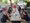 Eine Gruppe von Menschen sitzt an einem langen Tisch auf einer Außenterrasse und betrachtet Menükarten. Sie unterhalten sich in entspannter Atmosphäre. Im Hintergrund sind weitere Tische und ein weißes Auto zu erkennen.