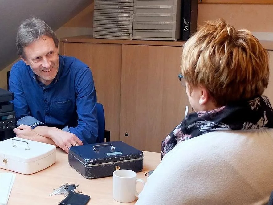 Im Büro: Ein Mann mit bleuem Hemd sitzt einer rotharigen Frau gegenüber und lächelt. Vor ihnen auf dem Tisch stehen zwei Geldkasetten und ein Schlüsselbund sowie eine Kaffetasse und einige Papiere.