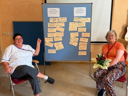 Zwei Frauen sitzen lächelnd vor einem blauen Board, das mit vielen gelben Notizzetteln bedeckt ist. Die Frau links hält einen Stift, die Frau rechts einen Blumenstrauß. An der Wand hängen Schilder mit Zukunftswünschen für Andrea und Maria.