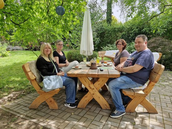 Vier Personen sitzen lächelnd an einem Holztisch im Garten und genießen Kaffee und Kuchen. Zwei Frauen sitzen links, rechts eine Frau und ein Mann. Im Hintergrund sind grüne Bäume und ein geschlossener weißer Sonnenschirm zu sehen.
