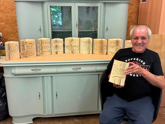 Ein lächelnder Mann hält einen gravierten Holzblock mit der Aufschrift „Welcome“. Auf dem hellblauen Schrank hinter ihm sind weitere Blöcke mit Inschriften wie „Home“ und „Love“ aufgereiht. Er präsentiert seine handgefertigten Werke.