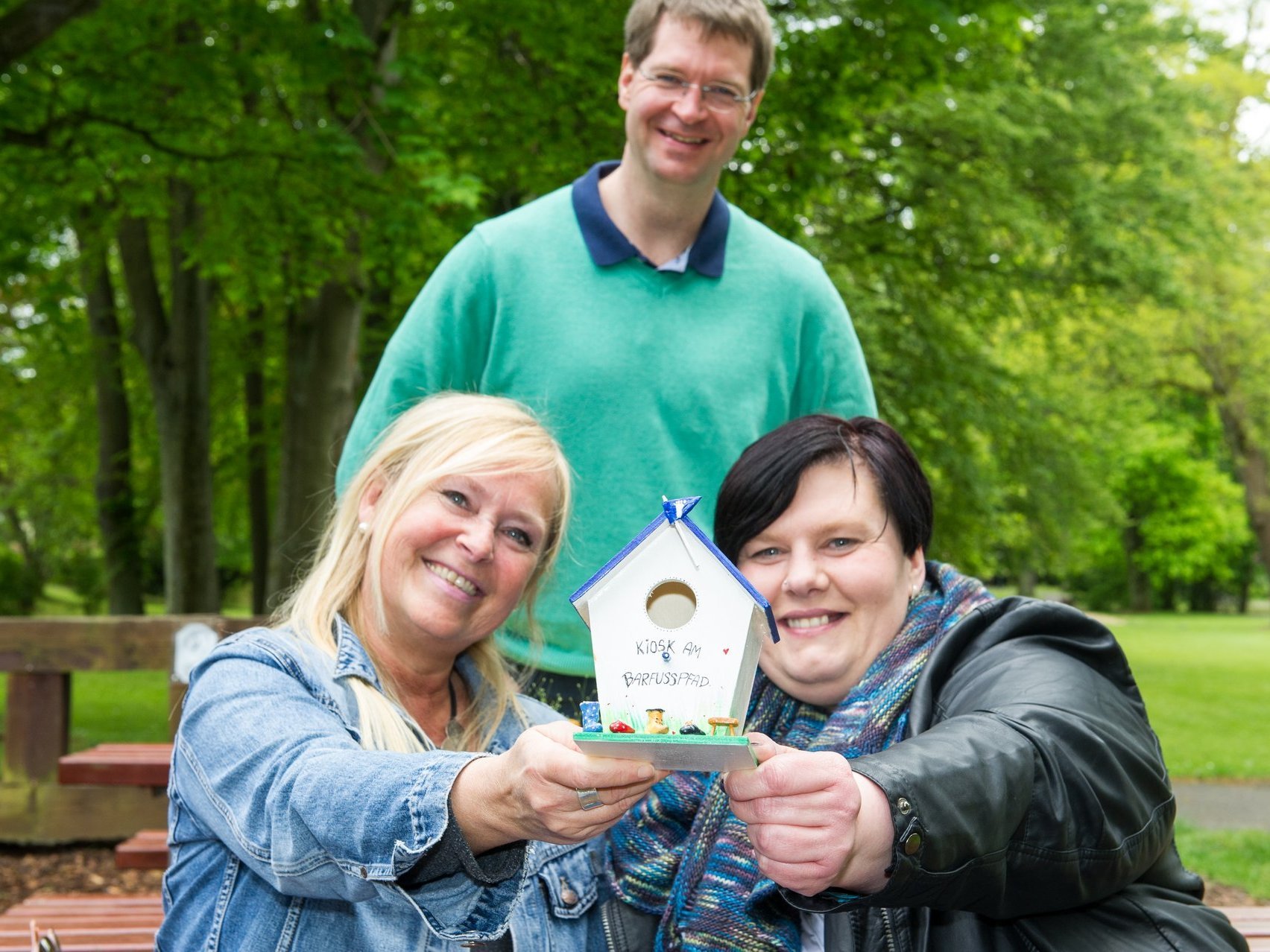 Zwei Frauen sitzen auf einer Parkbank und halten zusammen ein kleines Vogelhäuschen in der Hand.