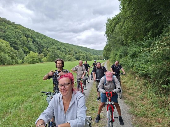 Eine Gruppe von Menschen radelt auf einem Feldweg durch eine grüne Landschaft mit bewaldeten Hügeln. Im Vordergrund lächelt eine Frau mit rosa Haaren in die Kamera, dahinter fahren weitere Radfahrer hinterher.