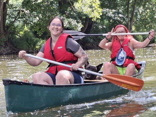 Zwei Frauen mit Brille und roten Schwimmwesten paddeln in einem grünen Kanu. Ein kleiner schwarzer Hund sitzt zwischen ihnen. Beide lächeln und halten Paddel auf einem Fluss, umgeben von Bäumen.