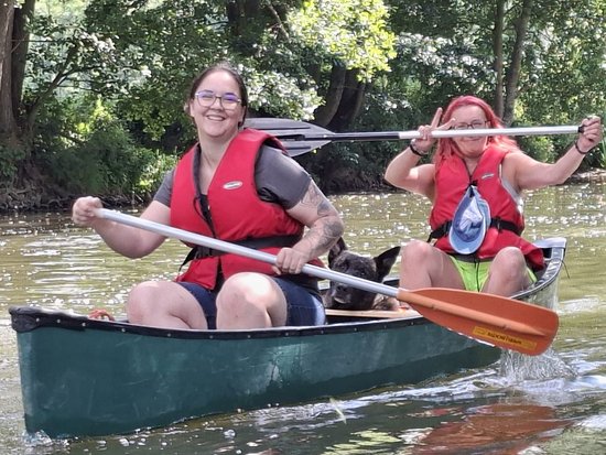 Zwei Frauen mit Brille und roten Schwimmwesten paddeln in einem grünen Kanu. Ein kleiner schwarzer Hund sitzt zwischen ihnen. Beide lächeln und halten Paddel auf einem Fluss, umgeben von Bäumen.