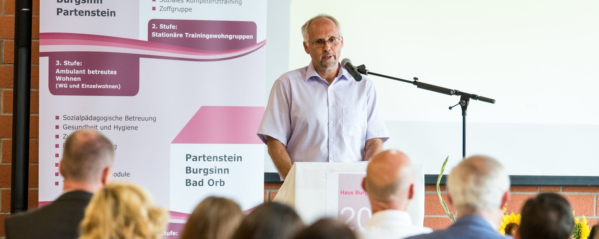 Ein Vortrag: Der Redner hat graues Haar, einen Kinnbart und trägt eine Brille. Im Hintergrund Plakate mit der Überschrift: "Bad Orb, Burgsinn, Partenstein"