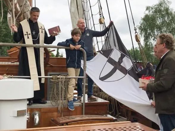 Pater Jörg hält eine Messe auf einem Segelboot - Helfer halten eine DO-Fahne