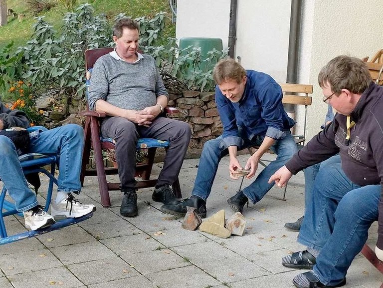 Vier Männer auf Gartenstühlen sitzen auf der Terasse und ordnen größere Steine auf dem Boden an